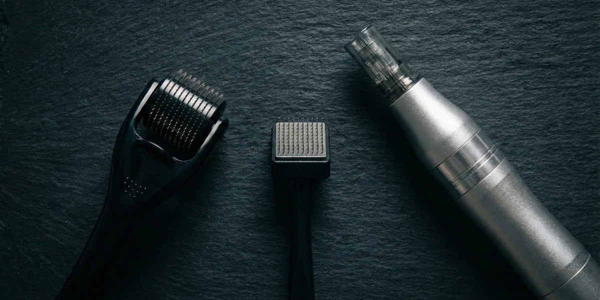 Three microneedling tools used for beard growth: a black derma roller, a square derma stamp, and a silver electric dermapen, arranged on a dark slate surface.