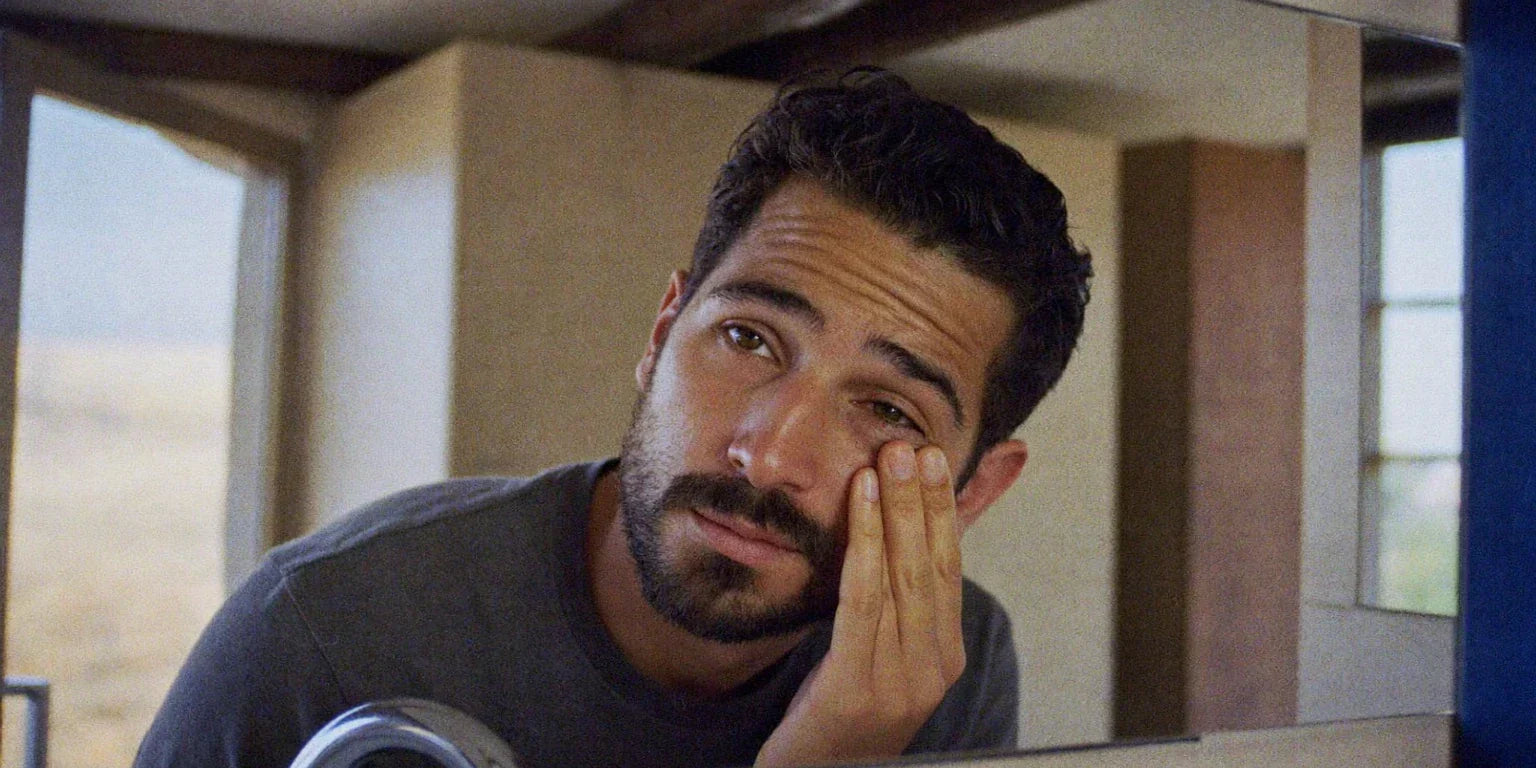 Tired-looking bearded man in his 30s rubbing the skin under his eye while leaning toward a mirror in warm natural light