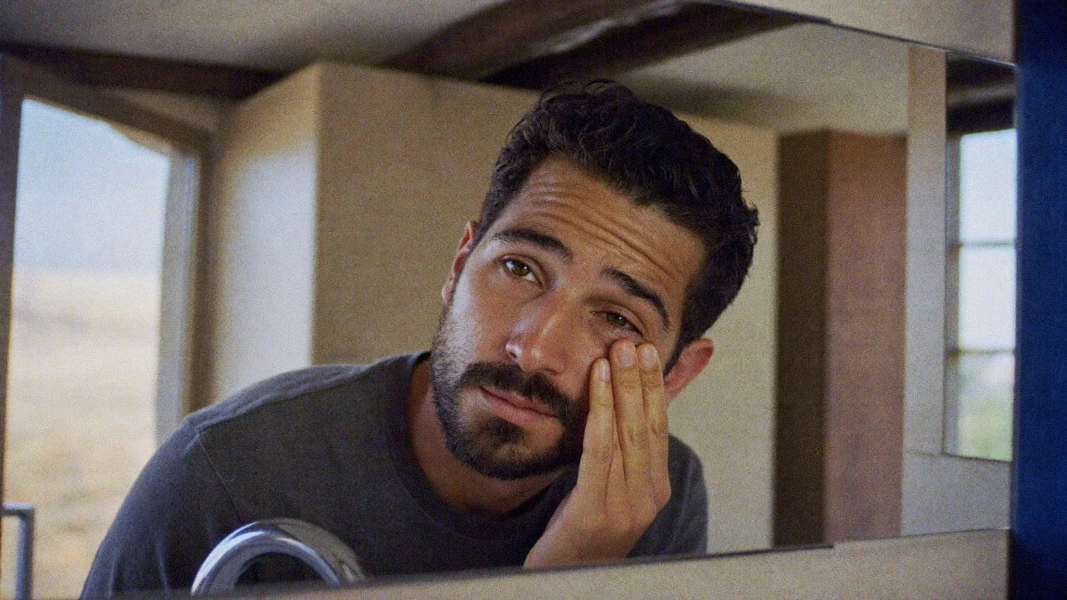 Tired-looking bearded man in his 30s rubbing the skin under his eye while leaning toward a mirror in warm natural light