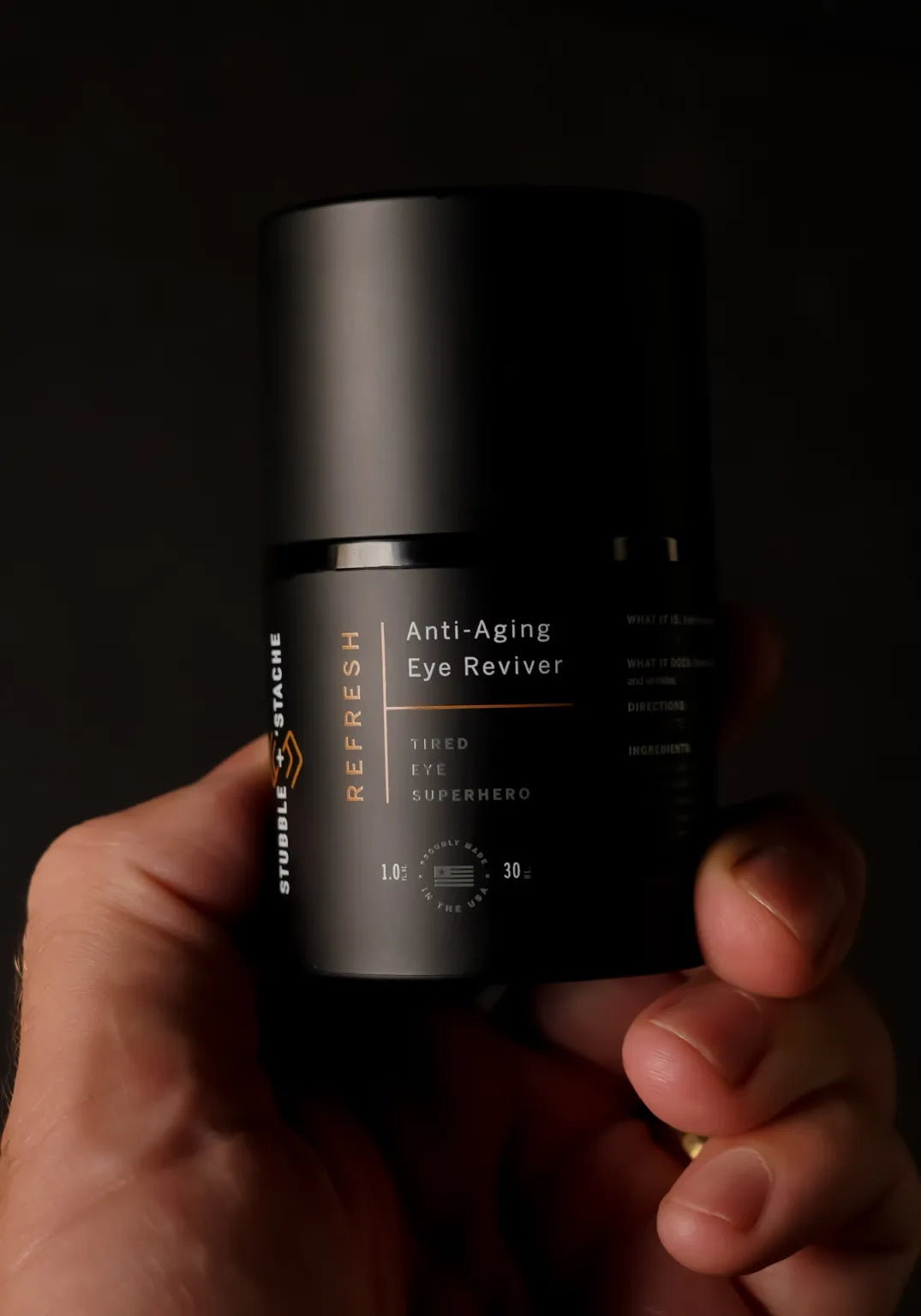 Hand holding Stubble & Stache Refresh Eye Cream with sleek black airless pump packaging against dark background
