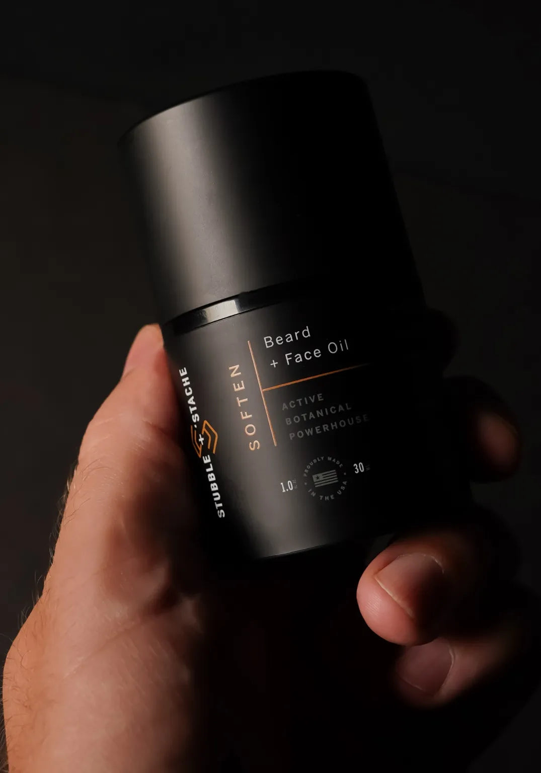 Hand holding Stubble & Stache Soften Face + Beard Oil in a sleek black airless pump bottle.