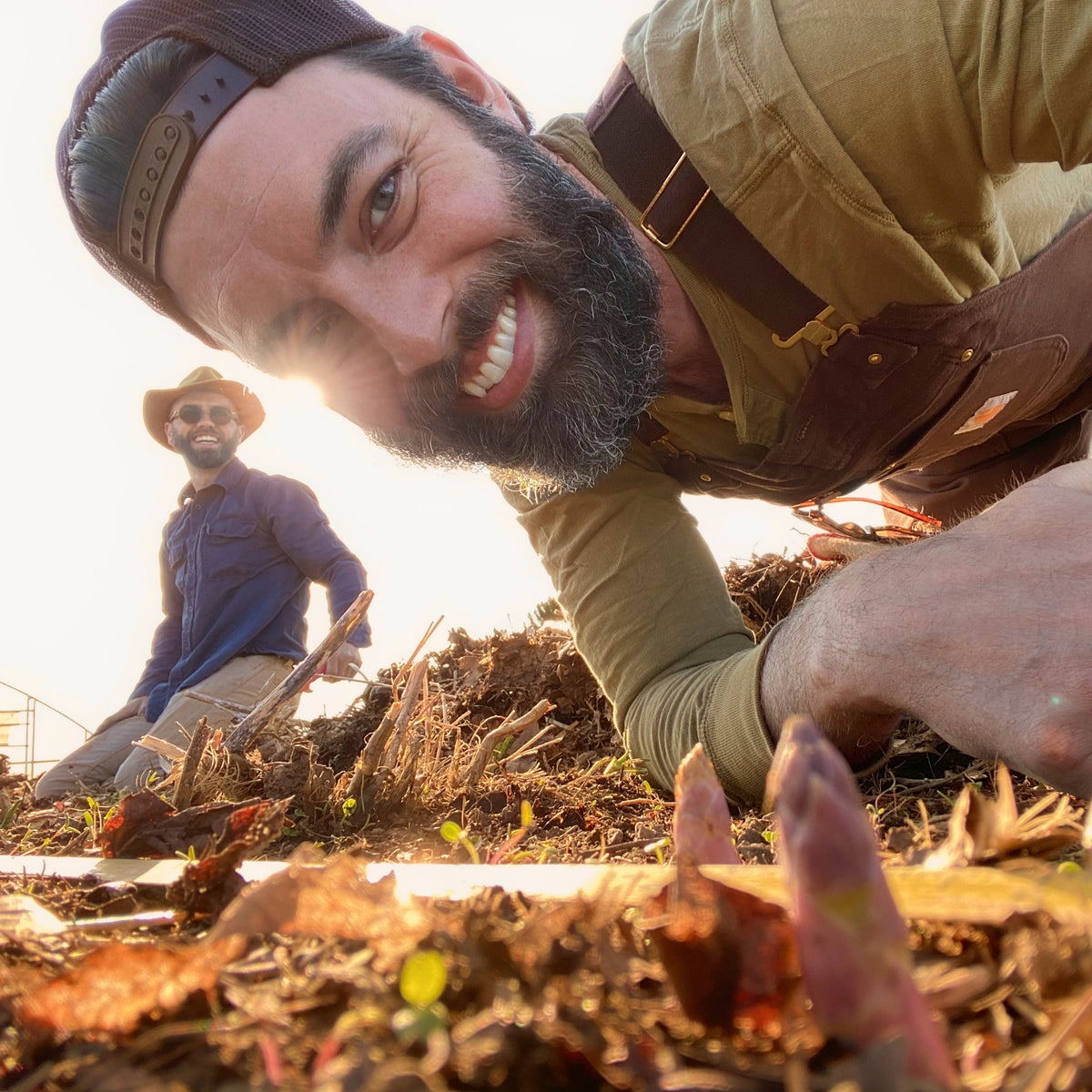 Nick Karnaze grinning at the camera from a garden bed at golden hour, a friend behind him