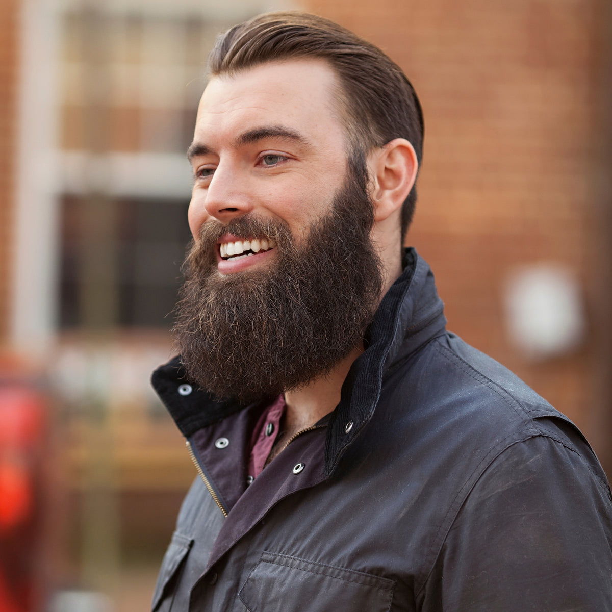 Nick Karnaze smiling in a waxed jacket outside a brick building, full beard