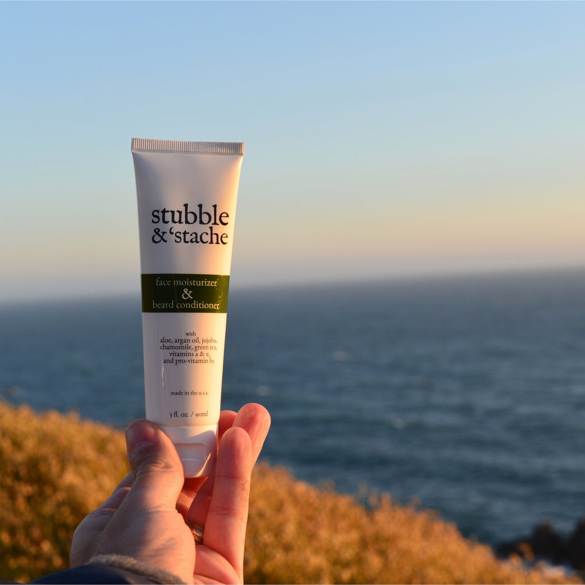 The original stubble & 'stache face moisturizer and beard conditioner tube held up at golden hour on the Northern California coast